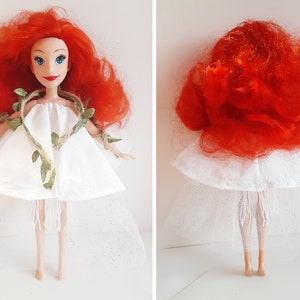 Puede incluir: Una mu&ntilde;eca de pelo rojo con un vestido blanco con un detalle de vid verde y una falda de tul blanca.