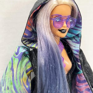 May include: A doll with lavender hair and purple sunglasses, wearing a black hooded jacket with a colorful, patterned lining. The jacket's sleeves feature a vibrant mix of green, blue, and purple designs. The doll has black lipstick and a purple top.