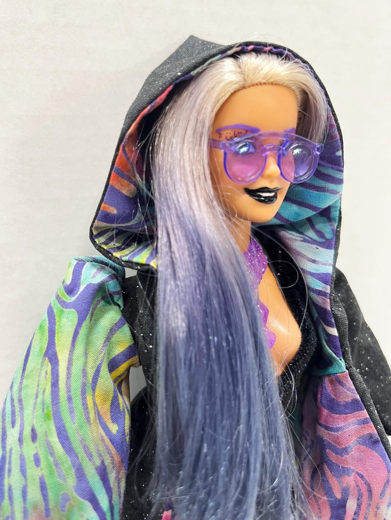 May include: A doll with lavender hair and purple sunglasses, wearing a black hooded jacket with a colorful, patterned lining. The jacket's sleeves feature a vibrant mix of green, blue, and purple designs. The doll has black lipstick and a purple top.