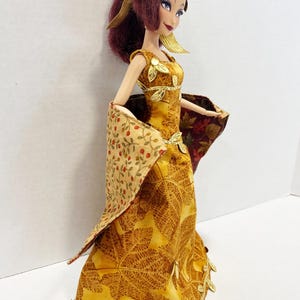 May include: A doll with dark red hair styled in a ponytail, wearing a floor-length yellow dress featuring a brown leaf pattern. The dress is accented with gold leaf embellishments at the neckline, waist, and hem, and includes a draped shawl.