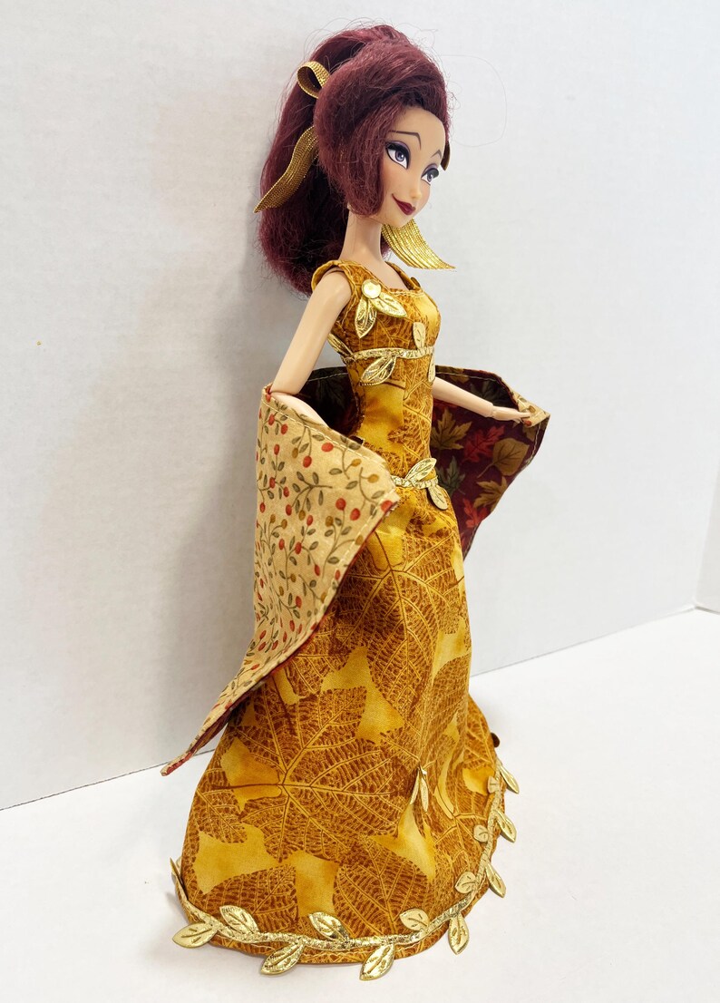 May include: A doll with dark red hair styled in a ponytail, wearing a floor-length yellow dress featuring a brown leaf pattern. The dress is accented with gold leaf embellishments at the neckline, waist, and hem, and includes a draped shawl.