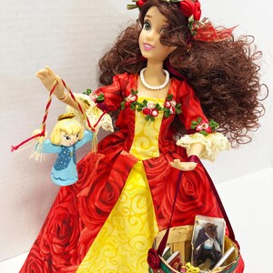 May include: A doll in a red rose-patterned dress with a yellow underskirt, holding a small basket and a miniature figure. The doll has brown hair, a red rose crown, and a pearl necklace. The basket contains miniature books and a picture.