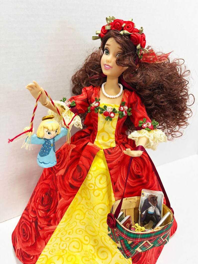 May include: A doll in a red rose-patterned dress with a yellow underskirt, holding a small basket and a miniature figure. The doll has brown hair, a red rose crown, and a pearl necklace. The basket contains miniature books and a picture.