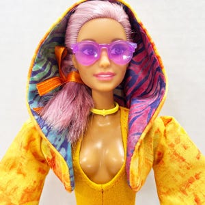 K&ouml;nnte beinhalten: Eine Puppe mit hellviolettem Haar tr&auml;gt eine durchscheinende violette Sonnenbrille, ein gelbes Halsband und einen gelben Body. Die Puppe tr&auml;gt eine Kapuzenjacke mit gelb-orangefarbenem Batikmuster au&szlig;en und einem bunten Innenfutter in Violett und Blau.