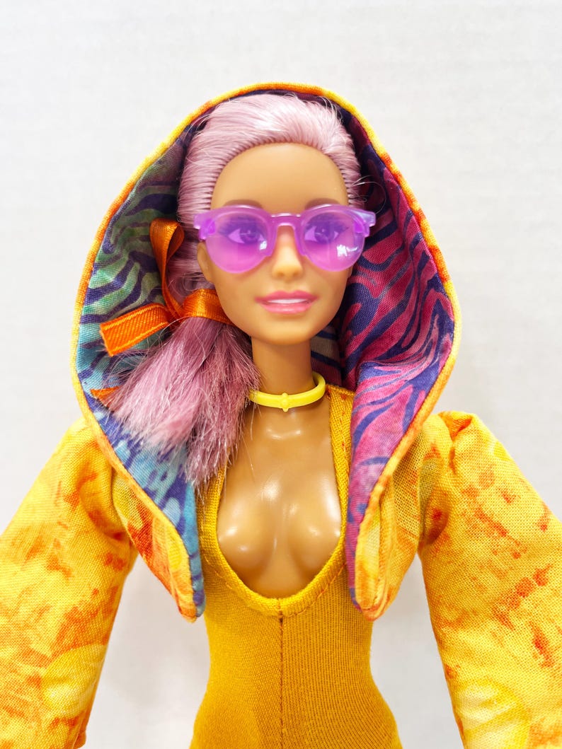 K&ouml;nnte beinhalten: Eine Puppe mit hellviolettem Haar tr&auml;gt eine durchscheinende violette Sonnenbrille, ein gelbes Halsband und einen gelben Body. Die Puppe tr&auml;gt eine Kapuzenjacke mit gelb-orangefarbenem Batikmuster au&szlig;en und einem bunten Innenfutter in Violett und Blau.