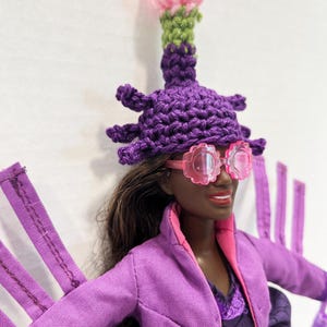 May include: A doll dressed in a purple outfit, pink-rimmed glasses, and a crocheted hat with a pink and green flower. The doll has dark skin and brown hair. The outfit includes a jacket and a dress.