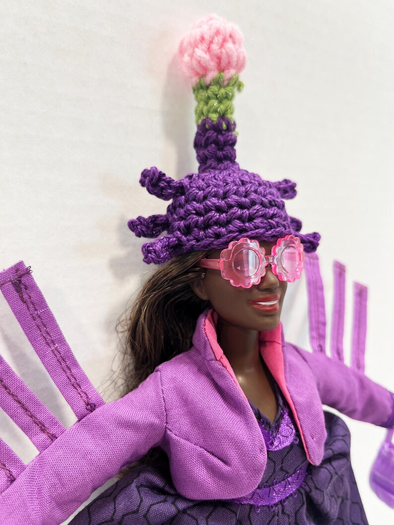 May include: A doll dressed in a purple outfit, pink-rimmed glasses, and a crocheted hat with a pink and green flower. The doll has dark skin and brown hair. The outfit includes a jacket and a dress.