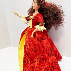 May include: A doll in a red rose-patterned gown with a yellow underskirt. The doll has long brown curly hair, a red rose crown, and a pearl necklace. The sleeves are trimmed with lace and small floral accents. The doll is a collectible item.