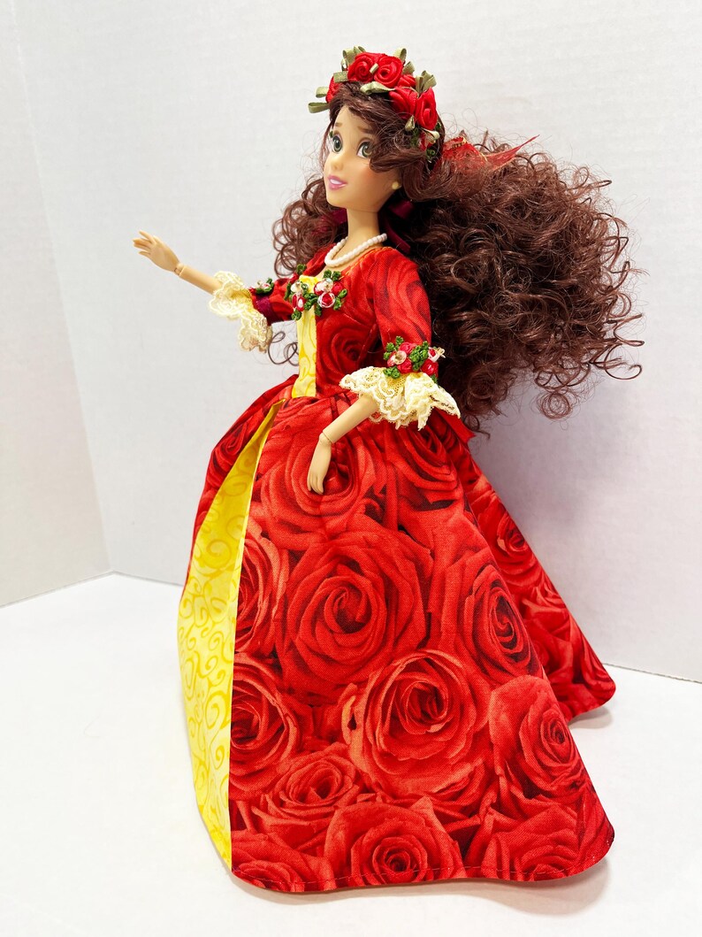 May include: A doll in a red rose-patterned gown with a yellow underskirt. The doll has long brown curly hair, a red rose crown, and a pearl necklace. The sleeves are trimmed with lace and small floral accents. The doll is a collectible item.