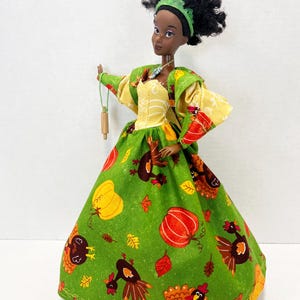 K&ouml;nnte beinhalten: Eine Puppe mit dunkler Haut und lockigem schwarzem Haar tr&auml;gt ein gr&uuml;nes Stirnband und ein Kleid mit einem Muster aus K&uuml;rbissen, Truth&auml;hnen und Herbstbl&auml;ttern. Das Kleid hat ein gelbes Oberteil und lange &Auml;rmel. Die Puppe h&auml;lt ein kleines Nudelholz.