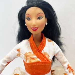 May include: A doll with long black hair and brown eyes wearing a white long-sleeved top featuring an orange leaf pattern. The garment has an orange wrap-style collar and a wide orange waistband with a white horizontal stripe. Small white stud earrings.