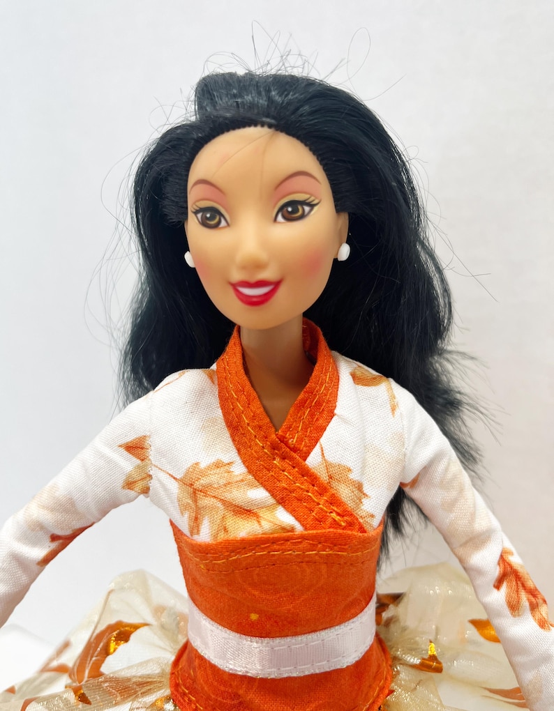 May include: A doll with long black hair and brown eyes wearing a white long-sleeved top featuring an orange leaf pattern. The garment has an orange wrap-style collar and a wide orange waistband with a white horizontal stripe. Small white stud earrings.