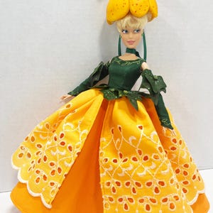 May include: A doll with blonde hair wearing a yellow headpiece resembling a flower, a dark green long-sleeved bodice with leaf-like accents, and a full-length yellow skirt featuring white eyelet embroidery and a scalloped hemline.