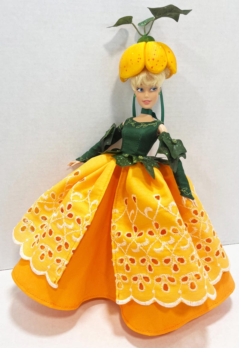 May include: A doll with blonde hair wearing a yellow headpiece resembling a flower, a dark green long-sleeved bodice with leaf-like accents, and a full-length yellow skirt featuring white eyelet embroidery and a scalloped hemline.
