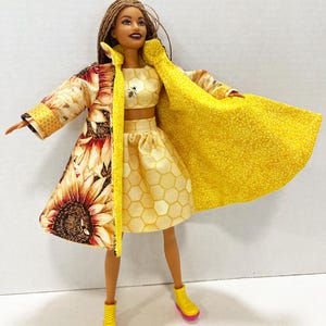Puede incluir: Una mu&ntilde;eca vestida con un conjunto de tres piezas. El atuendo incluye un top corto y una falda amarillos con estampado de panal, y un abrigo largo con un estampado de girasol en un lado y tela amarilla en el otro. La mu&ntilde;eca lleva botas amarillas con suelas rosas.