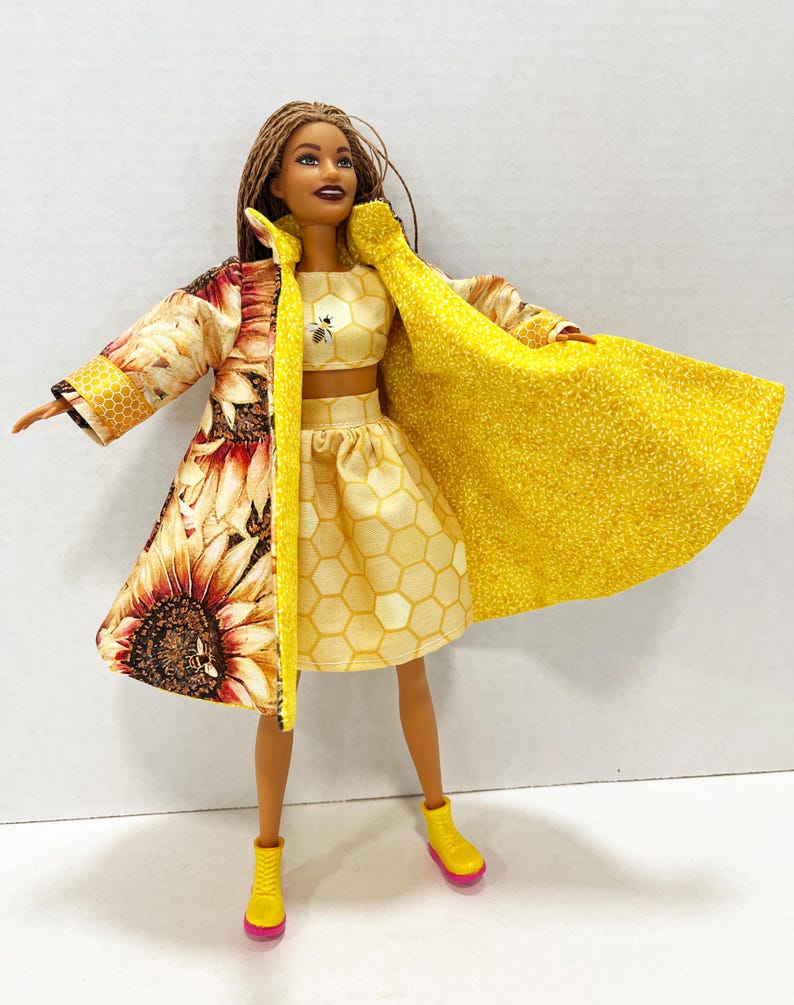 Puede incluir: Una mu&ntilde;eca vestida con un conjunto de tres piezas. El atuendo incluye un top corto y una falda amarillos con estampado de panal, y un abrigo largo con un estampado de girasol en un lado y tela amarilla en el otro. La mu&ntilde;eca lleva botas amarillas con suelas rosas.