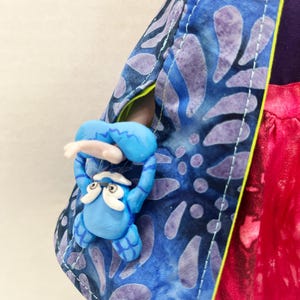 Pu&ograve; includere: Una bambola indossa un mantello blu con un motivo a foglie viola e una fodera verde lime brillante. Al mantello &egrave; fissata una piccola creatura blu scolpita con sopracciglia e barba bianche, posizionata come se fosse appesa al bordo.