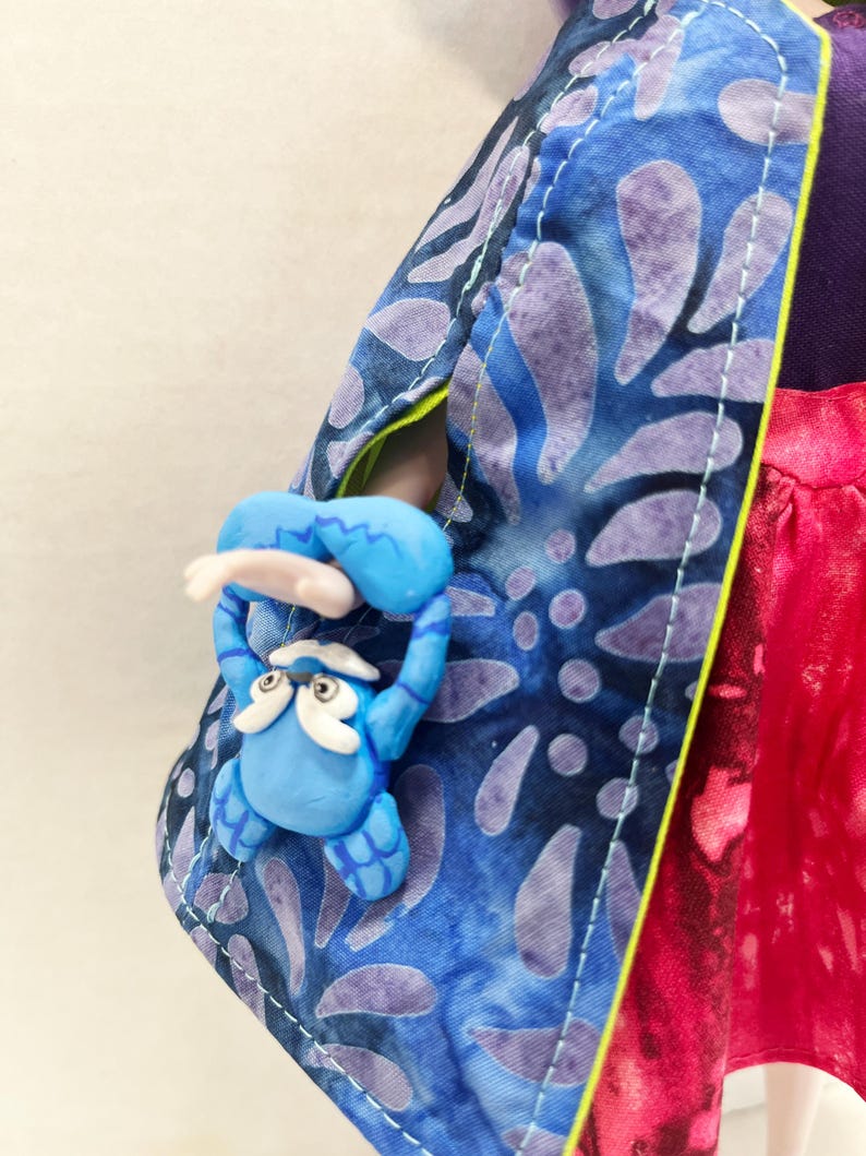 Pu&ograve; includere: Una bambola indossa un mantello blu con un motivo a foglie viola e una fodera verde lime brillante. Al mantello &egrave; fissata una piccola creatura blu scolpita con sopracciglia e barba bianche, posizionata come se fosse appesa al bordo.