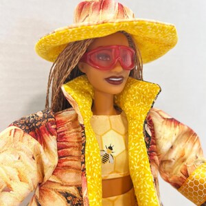 Puede incluir: Una mu&ntilde;eca vestida con un conjunto amarillo y con estampado floral, que incluye un sombrero, una chaqueta y un top corto. El top corto presenta un gr&aacute;fico de abeja. La mu&ntilde;eca tambi&eacute;n lleva gafas tintadas de rojo.