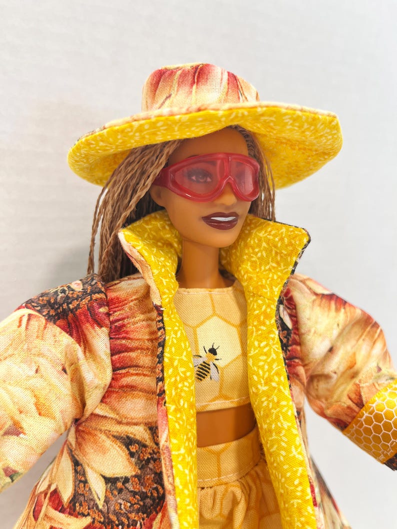 Puede incluir: Una mu&ntilde;eca vestida con un conjunto amarillo y con estampado floral, que incluye un sombrero, una chaqueta y un top corto. El top corto presenta un gr&aacute;fico de abeja. La mu&ntilde;eca tambi&eacute;n lleva gafas tintadas de rojo.