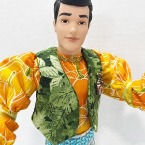 以下が含まれることがあります： カボチャ柄のオレンジ色のシャツの上に緑色のベストを着た人形。人形は黒髪で、星柄の青いズボンを履いています。金のネックレスとブロンズのカボチャのピンも見えます。