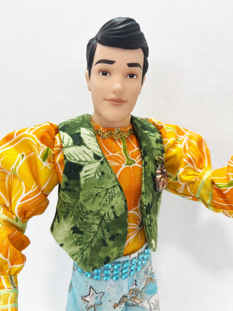以下が含まれることがあります： カボチャ柄のオレンジ色のシャツの上に緑色のベストを着た人形。人形は黒髪で、星柄の青いズボンを履いています。金のネックレスとブロンズのカボチャのピンも見えます。