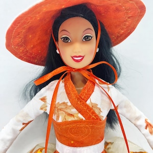 May include: A doll with long black hair wearing a wide-brimmed orange hat with a matching ribbon tie. The doll is dressed in a white long-sleeved top featuring an orange leaf pattern, an orange bodice, and a white skirt with orange leaf accents.