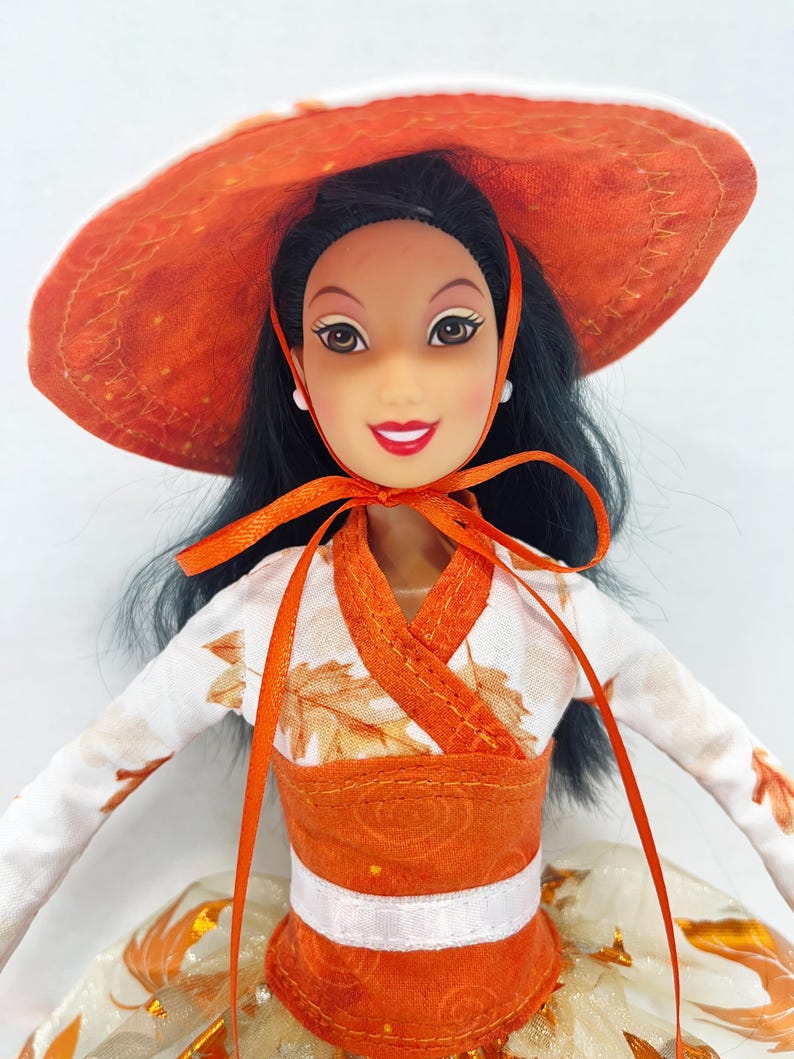 May include: A doll with long black hair wearing a wide-brimmed orange hat with a matching ribbon tie. The doll is dressed in a white long-sleeved top featuring an orange leaf pattern, an orange bodice, and a white skirt with orange leaf accents.