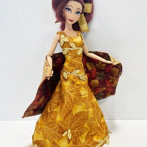 May include: A doll with dark red hair styled in a ponytail with a gold ribbon. The doll wears a floor-length yellow dress featuring a brown leaf pattern, accented with gold leaf trim at the neckline, waist, and hem, and a matching cape with a dark red leaf print.