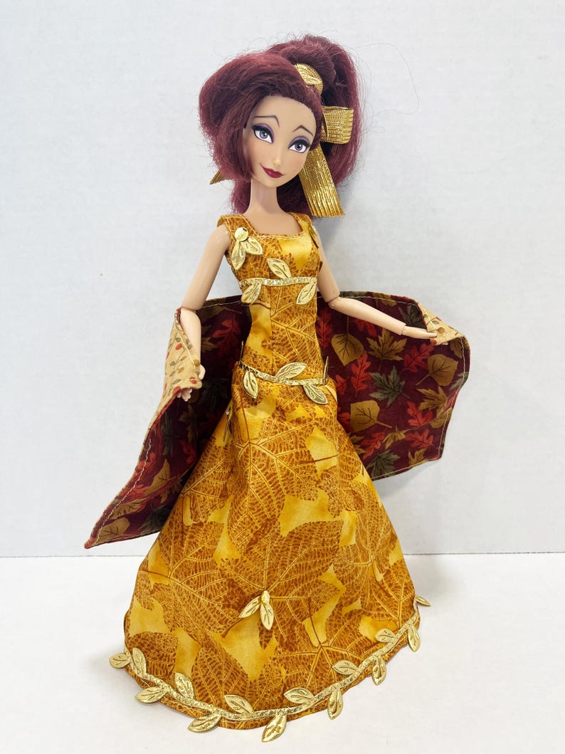 May include: A doll with dark red hair styled in a ponytail with a gold ribbon. The doll wears a floor-length yellow dress featuring a brown leaf pattern, accented with gold leaf trim at the neckline, waist, and hem, and a matching cape with a dark red leaf print.