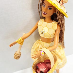 Puede incluir: Mu&ntilde;eca vestida con un atuendo amarillo con estampado de panal, con un sombrero y pu&ntilde;os a juego, sosteniendo una cesta con un animal peque&ntilde;o. Lleva un cuchar&oacute;n de miel de madera. El atuendo de la mu&ntilde;eca tiene una abeja bordada.