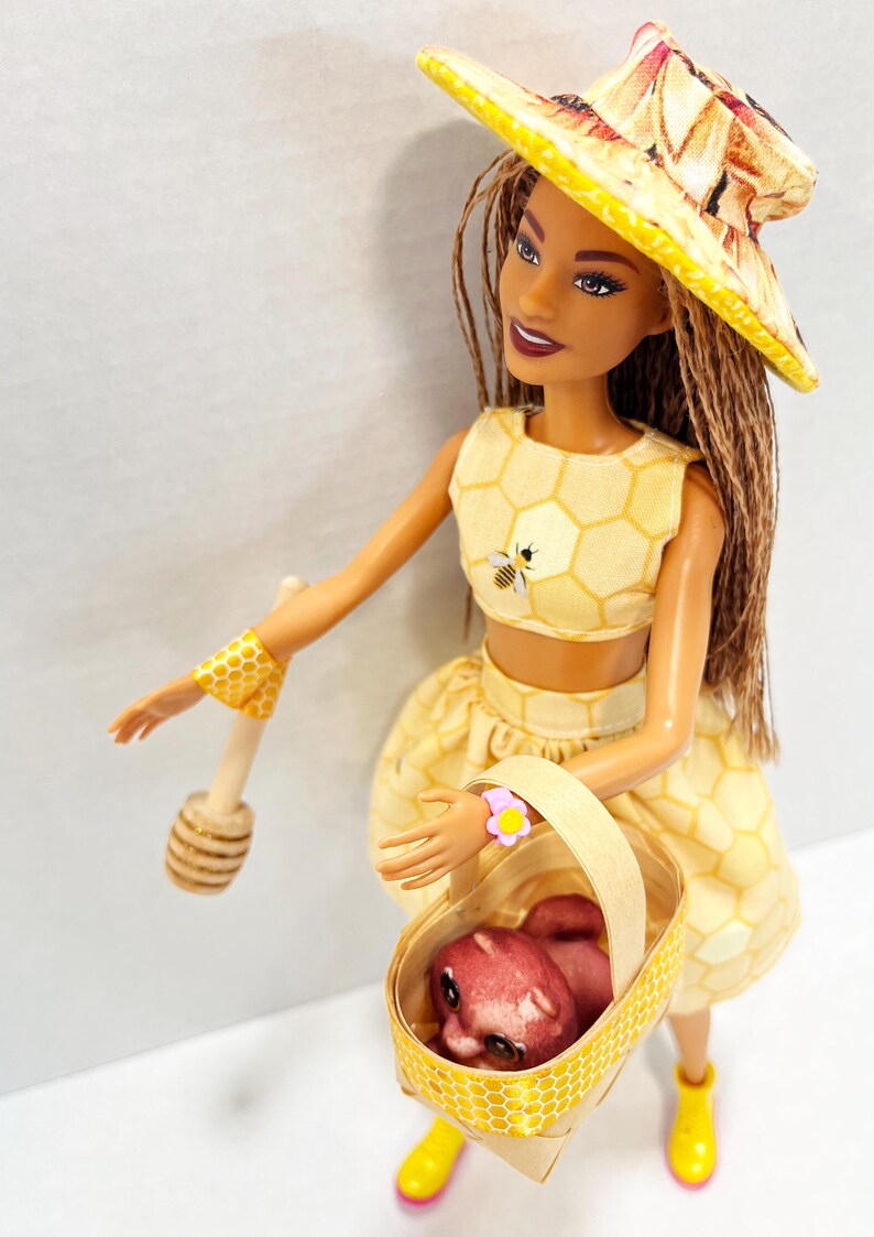 Puede incluir: Mu&ntilde;eca vestida con un atuendo amarillo con estampado de panal, con un sombrero y pu&ntilde;os a juego, sosteniendo una cesta con un animal peque&ntilde;o. Lleva un cuchar&oacute;n de miel de madera. El atuendo de la mu&ntilde;eca tiene una abeja bordada.