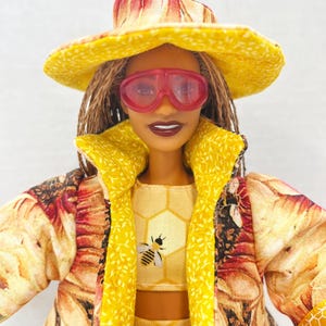 Puede incluir: Una mu&ntilde;eca vestida con un conjunto amarillo con un motivo de abeja, un sombrero a juego y gafas de sol rosas. El conjunto incluye un top corto, una falda y un abrigo largo con estampado floral. La mu&ntilde;eca tiene el pelo casta&ntilde;o y l&aacute;piz labial oscuro.