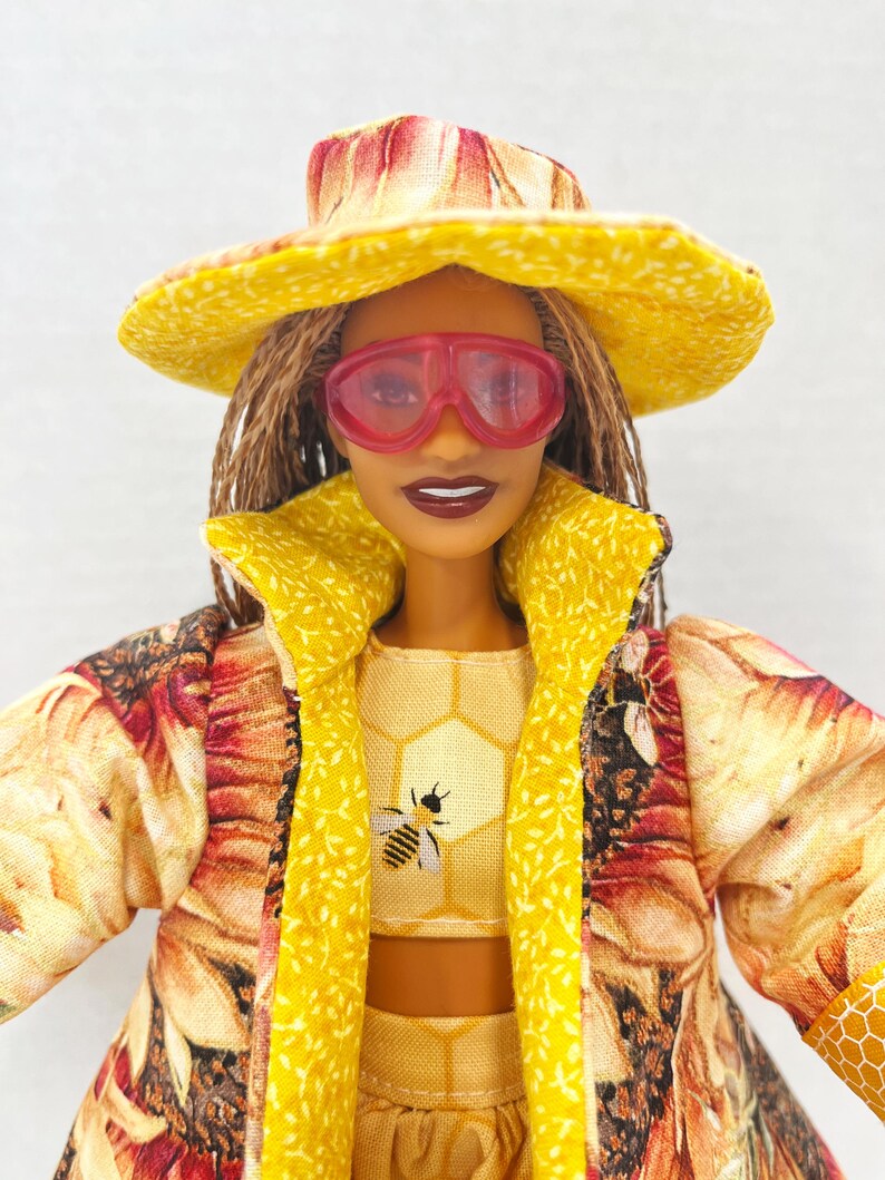 Puede incluir: Una mu&ntilde;eca vestida con un conjunto amarillo con un motivo de abeja, un sombrero a juego y gafas de sol rosas. El conjunto incluye un top corto, una falda y un abrigo largo con estampado floral. La mu&ntilde;eca tiene el pelo casta&ntilde;o y l&aacute;piz labial oscuro.