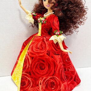 May include: A doll in a red rose-print gown with yellow accents. The doll has brown curly hair, a floral crown, and a pearl necklace. The sleeves have lace trim and floral accents. The doll is posed with one arm raised.