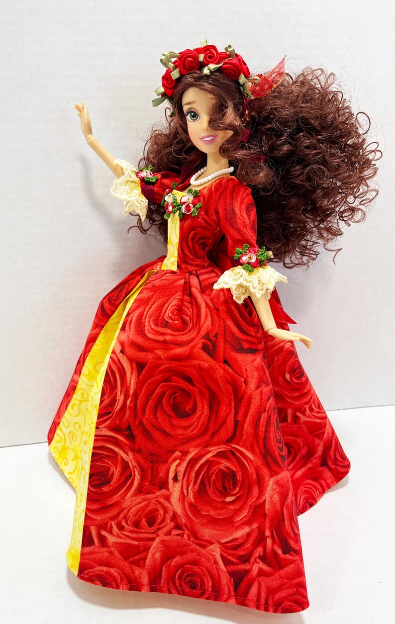May include: A doll in a red rose-print gown with yellow accents. The doll has brown curly hair, a floral crown, and a pearl necklace. The sleeves have lace trim and floral accents. The doll is posed with one arm raised.