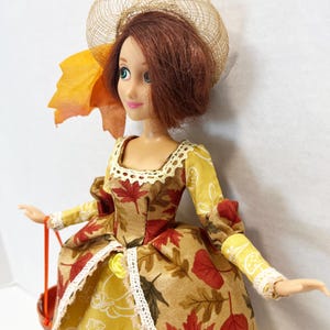 May include: A doll with auburn hair wearing a tan hat and a dress featuring a pattern of red and brown autumn leaves, yellow pumpkins, and acorns. The dress has long sleeves, a lace-trimmed neckline, and a lace-trimmed skirt overlay.