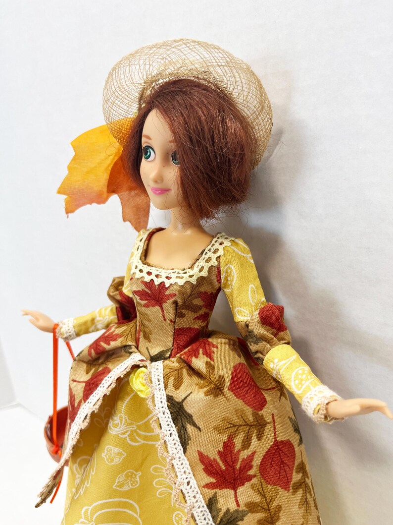 May include: A doll with auburn hair wearing a tan hat and a dress featuring a pattern of red and brown autumn leaves, yellow pumpkins, and acorns. The dress has long sleeves, a lace-trimmed neckline, and a lace-trimmed skirt overlay.
