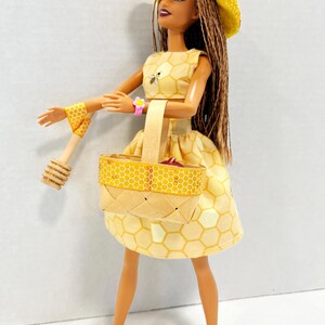 Puede incluir: Mu&ntilde;eca vestida con un conjunto amarillo con estampado de panal, con sombrero y cesta a juego. La mu&ntilde;eca lleva una pulsera rosa y botas amarillas con suelas rosas. Un cuchar&oacute;n de miel de madera est&aacute; en su mano.
