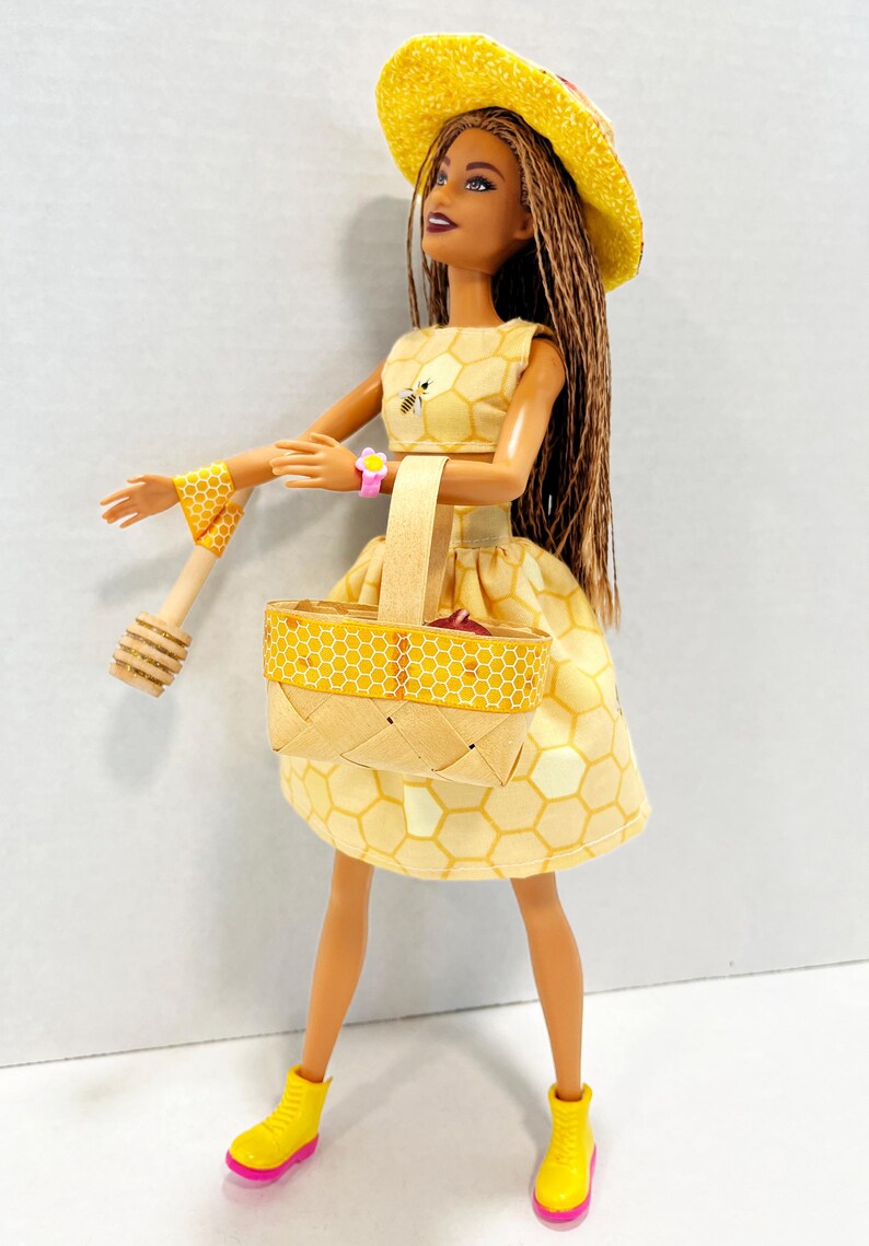 Puede incluir: Mu&ntilde;eca vestida con un conjunto amarillo con estampado de panal, con sombrero y cesta a juego. La mu&ntilde;eca lleva una pulsera rosa y botas amarillas con suelas rosas. Un cuchar&oacute;n de miel de madera est&aacute; en su mano.