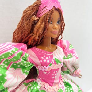 May include: A doll with brown hair and a pink tiara. The doll wears a pink, white, and green patterned dress with puff sleeves and a sequined bodice. The doll's skin tone is brown.