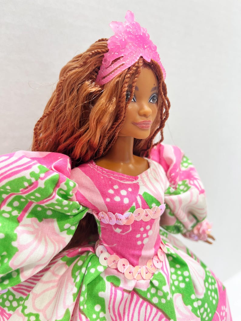 May include: A doll with brown hair and a pink tiara. The doll wears a pink, white, and green patterned dress with puff sleeves and a sequined bodice. The doll's skin tone is brown.