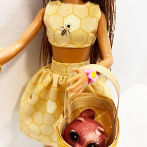 Puede incluir: Una mu&ntilde;eca con un conjunto amarillo con estampado de panal, que incluye un top corto con una abeja y una falda a juego. La mu&ntilde;eca lleva una cesta con una criatura roja. La cesta tiene un dise&ntilde;o de panal amarillo. La mu&ntilde;eca tiene pelo casta&ntilde;o y un sombrero amarillo.