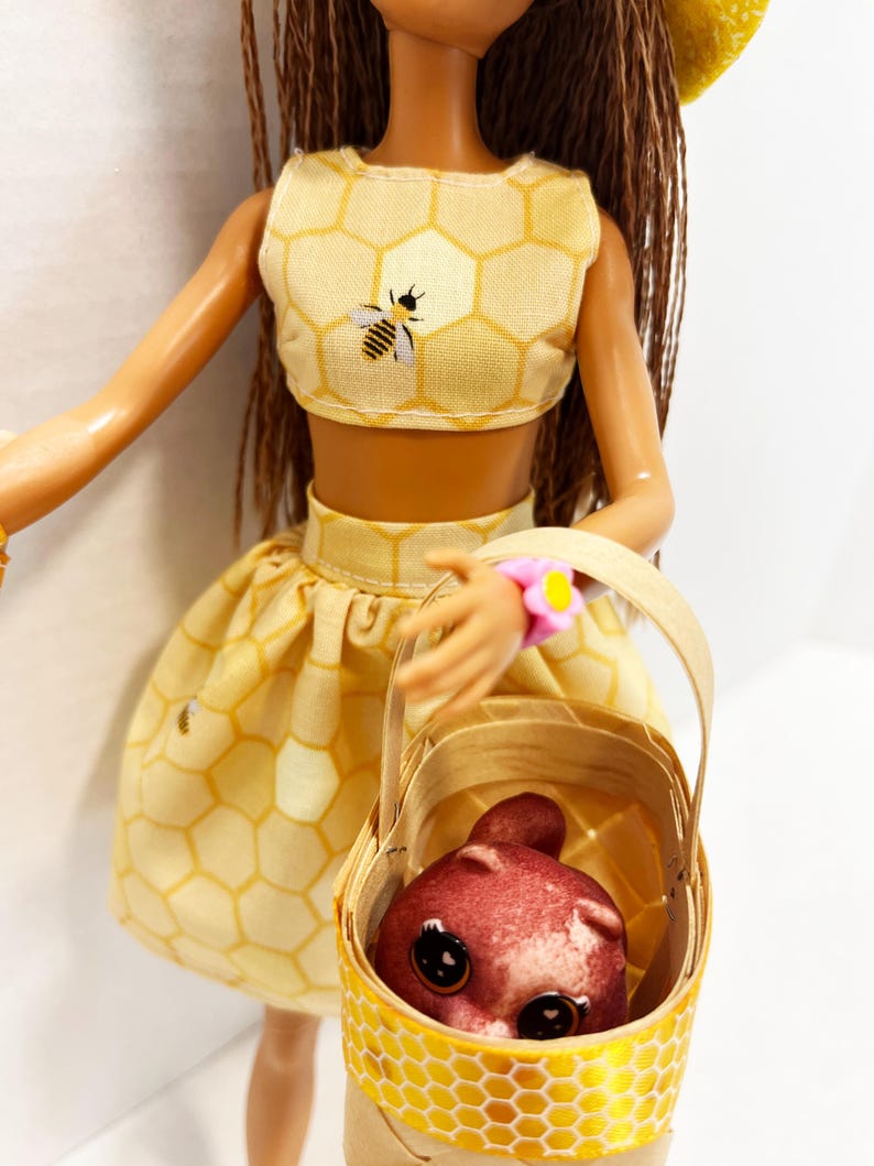 Puede incluir: Una mu&ntilde;eca con un conjunto amarillo con estampado de panal, que incluye un top corto con una abeja y una falda a juego. La mu&ntilde;eca lleva una cesta con una criatura roja. La cesta tiene un dise&ntilde;o de panal amarillo. La mu&ntilde;eca tiene pelo casta&ntilde;o y un sombrero amarillo.
