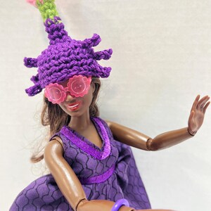 May include: A doll dressed in a purple outfit, including a crochet hat with spiky details and pink flower accents, and pink flower-shaped sunglasses. The doll also wears a purple dress and bracelet.