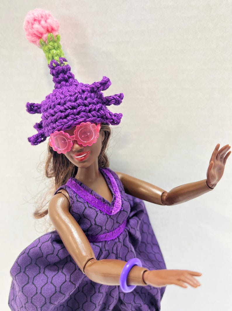 May include: A doll dressed in a purple outfit, including a crochet hat with spiky details and pink flower accents, and pink flower-shaped sunglasses. The doll also wears a purple dress and bracelet.
