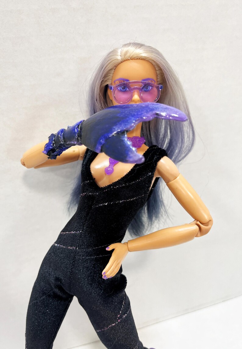 May include: A doll in a black jumpsuit with silver accents, purple-tinted sunglasses, and a large, dark blue claw-like accessory. The doll has lavender hair and a matching purple necklace. The overall aesthetic is futuristic and stylish.
