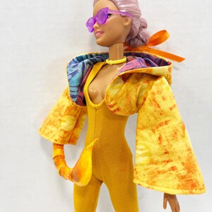 K&ouml;nnte beinhalten: Eine Puppe mit hellrosa Haar in einem Dutt, tr&auml;gt eine violette Sonnenbrille, ein gelbes Halsband und einen senfgelben Body. Die Puppe tr&auml;gt eine gelb-orange gemusterte Kapuzenjacke mit weiten &Auml;rmeln und ein passendes, geschwungenes Schwanz-Accessoire.