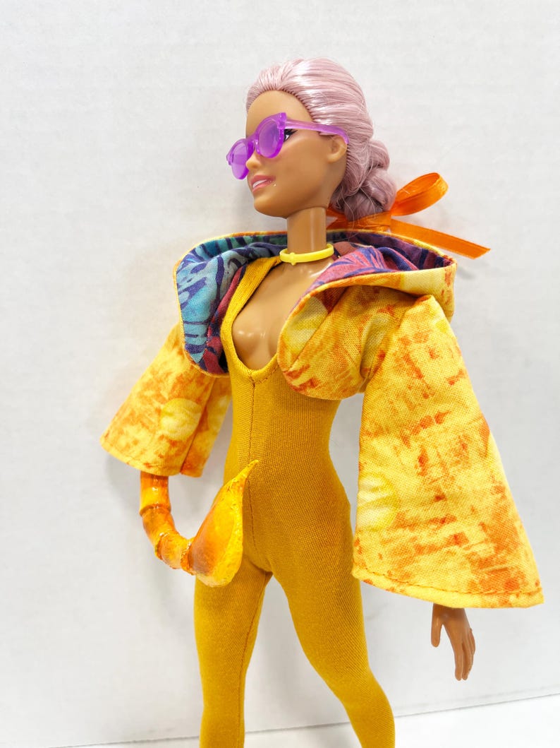 K&ouml;nnte beinhalten: Eine Puppe mit hellrosa Haar in einem Dutt, tr&auml;gt eine violette Sonnenbrille, ein gelbes Halsband und einen senfgelben Body. Die Puppe tr&auml;gt eine gelb-orange gemusterte Kapuzenjacke mit weiten &Auml;rmeln und ein passendes, geschwungenes Schwanz-Accessoire.