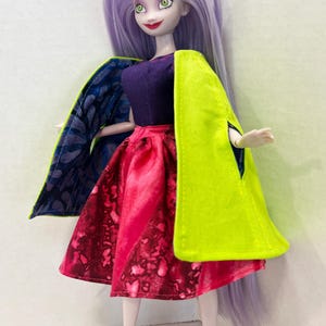 Pu&ograve; includere: Una bambola con lunghi capelli color lavanda, occhi verdi e labbra rosse. La bambola indossa un top viola scuro, una gonna con motivo rosso e rosa, un mantello verde lime brillante con fodera a motivi blu e viola e tacchi viola.